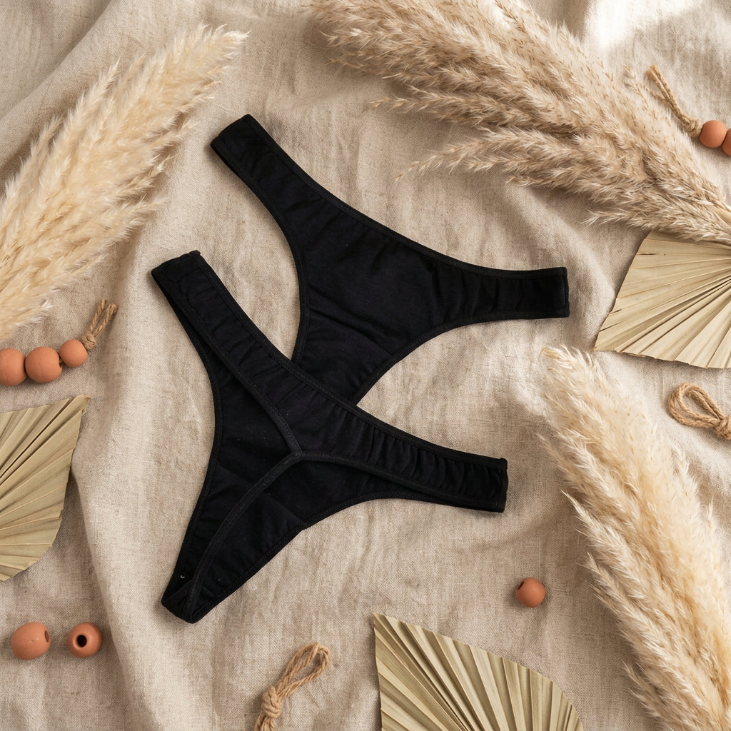 Black thong-style underwear laid flat on a light gray fabric surface, with a white floral card partially visible in the top left corner.