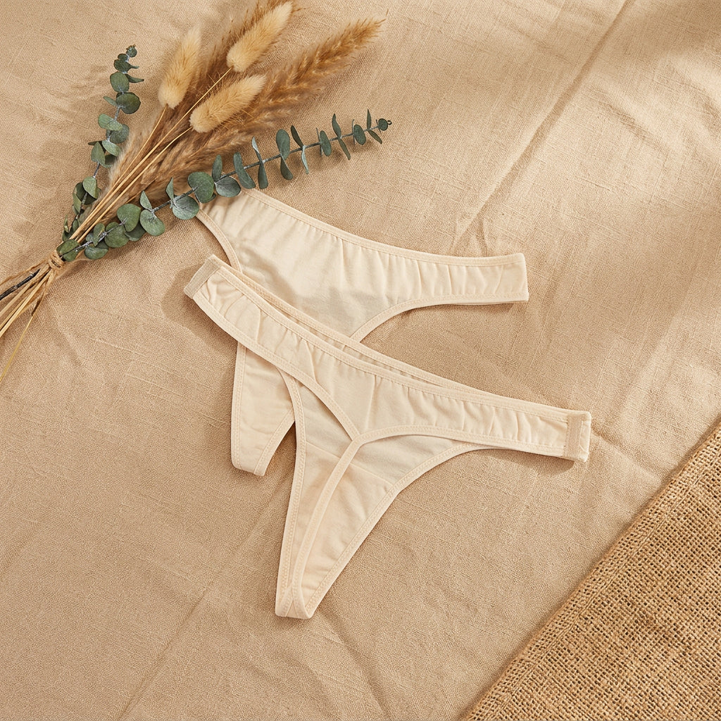 Two pairs of light beige thong underwear laid flat on a gray fabric surface, with a decorative card and cotton stems in the top left corner.