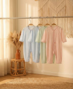 Three baby rompers in light blue, green, and pink hanging on a rack with a decorative wall and stool in the background.
