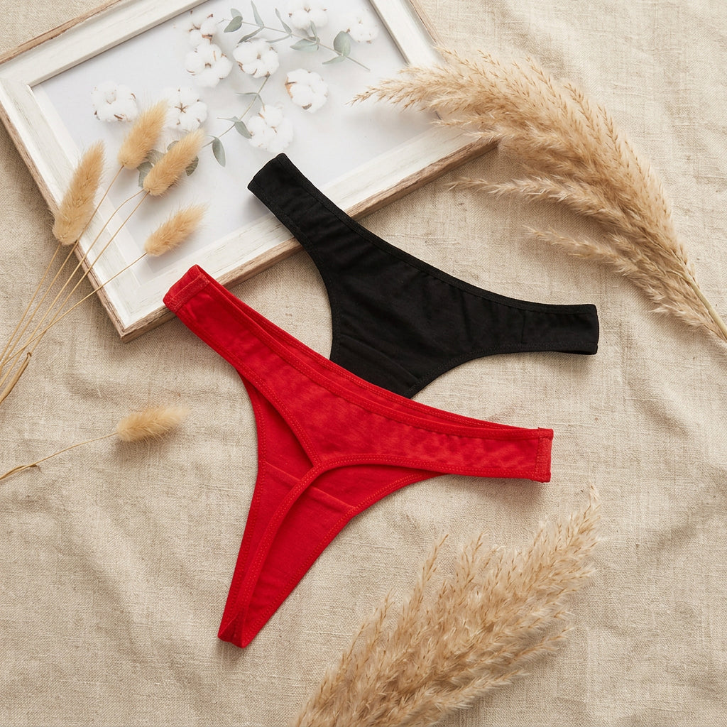 Two pairs of thong-style underwear laid flat on a light gray fabric surface: one bright red pair in front and one black pair behind, with a white floral print partially visible in the top left corner.