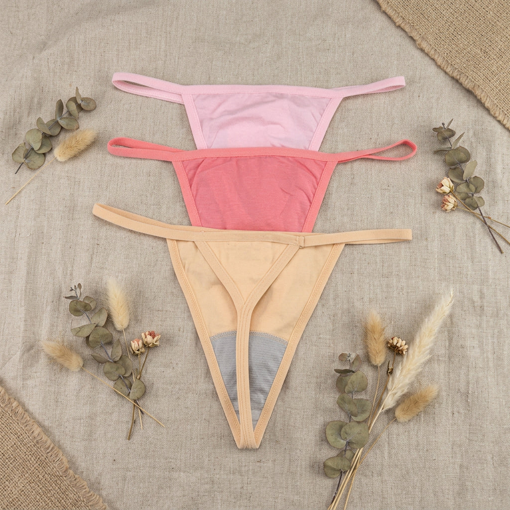 Three pairs of women's thong-style underwear laid flat on a gray background, stacked vertically in light pink, coral pink, and pale yellow, with a small gray panel at the front of the bottom pair.