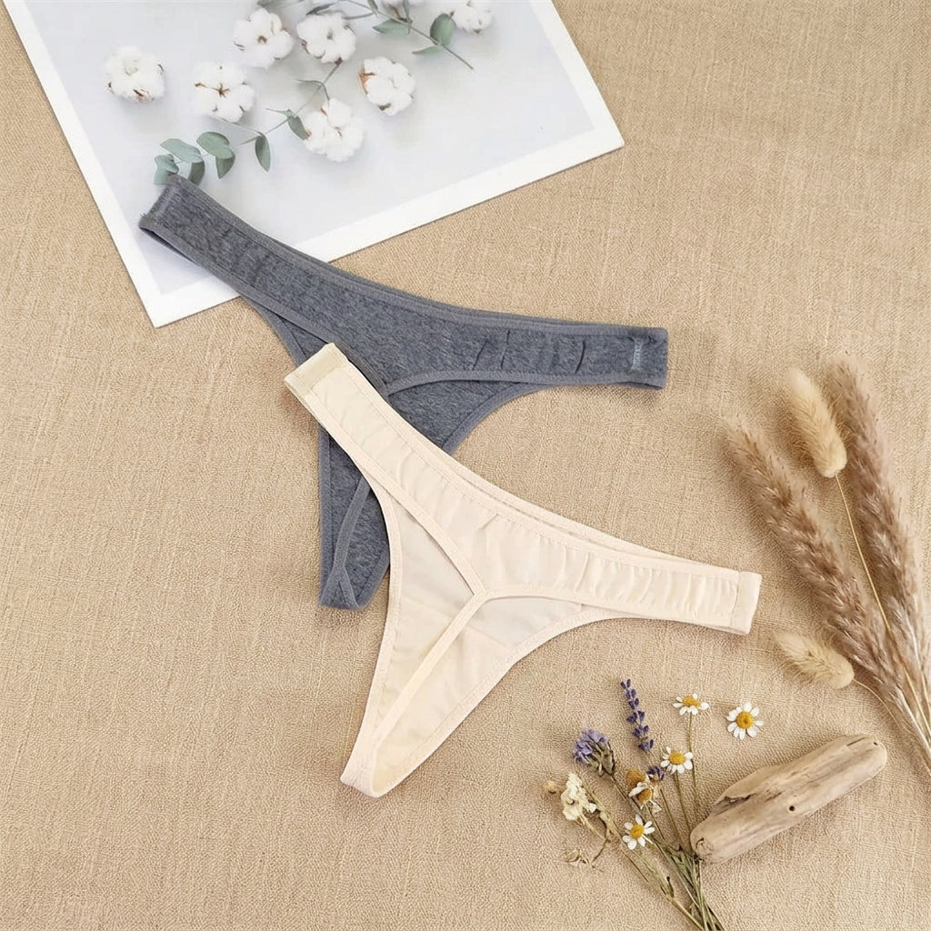 Two pairs of thong-style underwear laid flat on a light gray fabric surface: one dark gray and one light cream. A white card with small white flowers rests in the upper left corner as a styling prop.