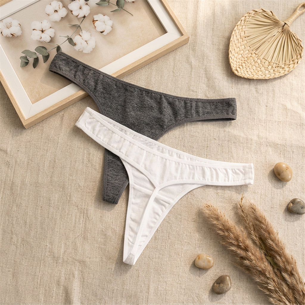 Two pairs of thong-style underwear laid flat on a gray fabric surface: one white pair in front and one dark heather gray pair behind, with a white card and cotton stems as styling props.