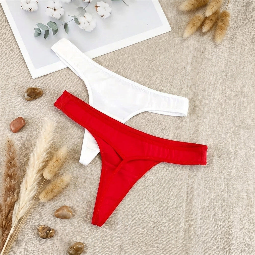 Two pairs of thong-style underwear laid flat on a light gray fabric surface: one white pair and one bright red pair, with a small decorative card and cotton stems in the top left corner.